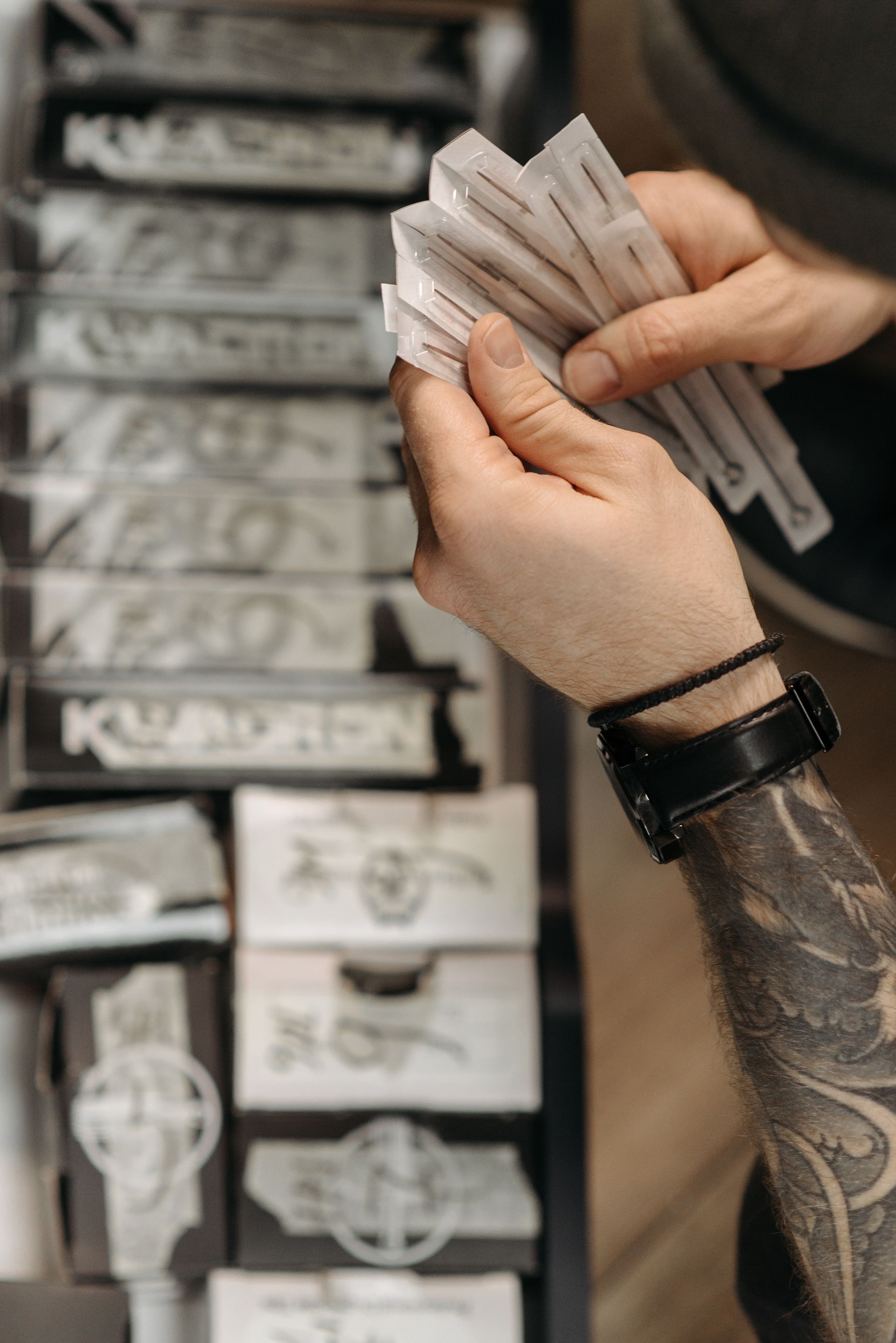 A tattoo artist’s hand holds several individually packaged needle cartridges in front of shelves containing tattoo supplies.