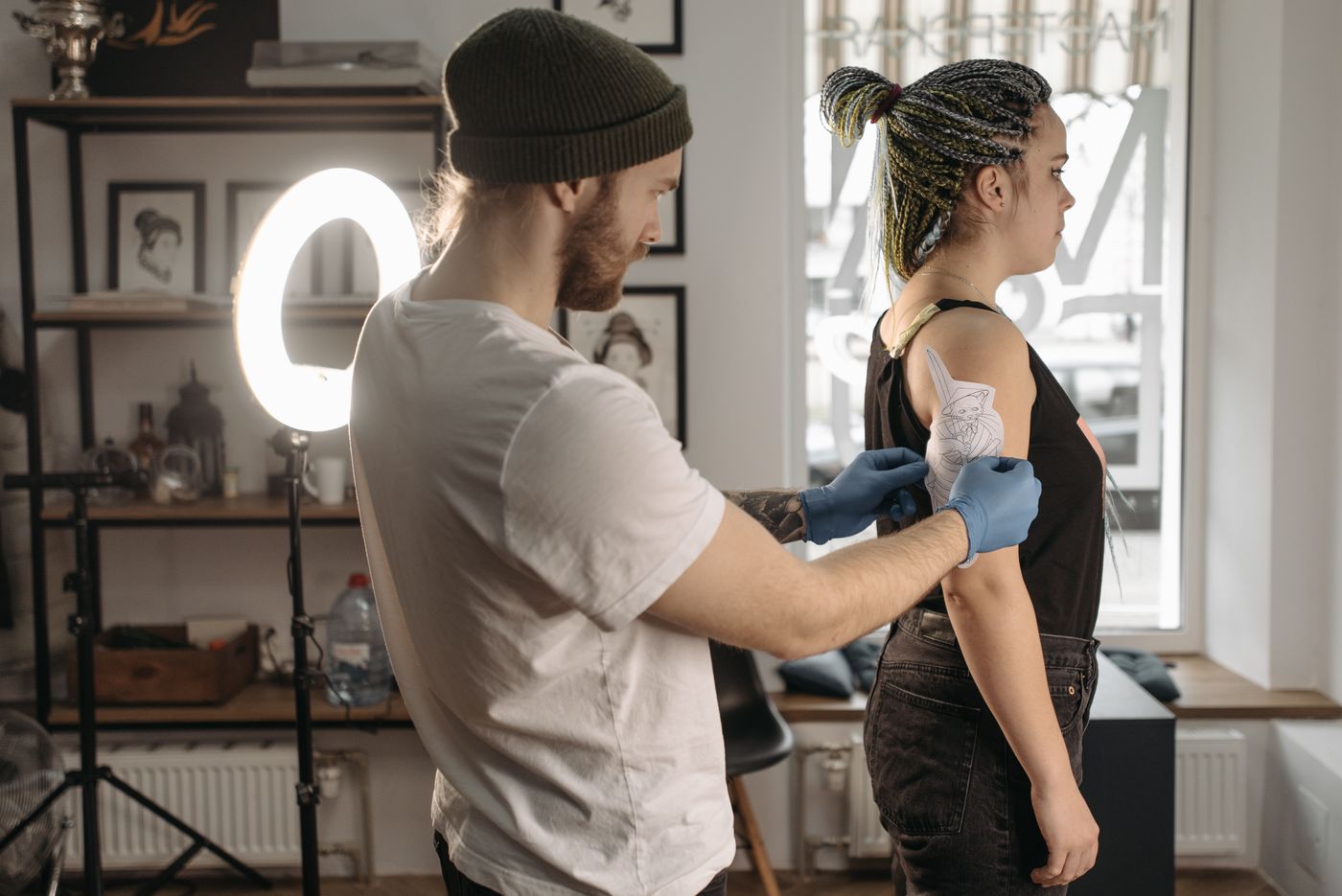 A tattoo artist wearing blue gloves positions an outlined tattoo stencil on a woman's upper arm in a studio.