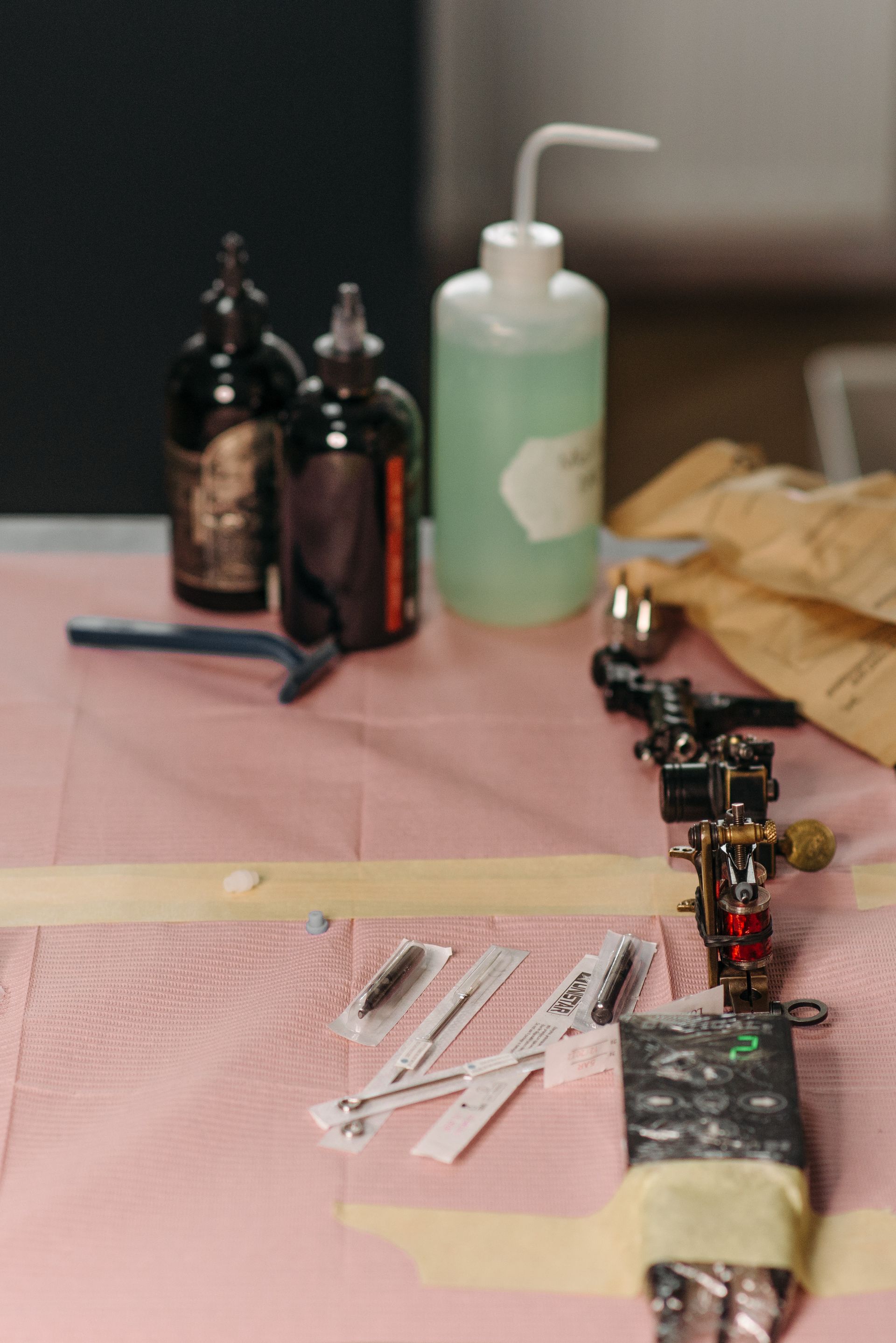 A tattoo workstation displays ink bottles, a green cleaning-solution bottle, packaged needles, a tattoo machine, and other supplies on a covered surface.