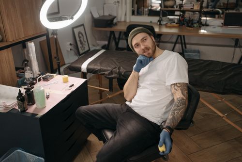 Free Tattoo artist sitting in a studio with equipment, wearing gloves and a beanie. Stock Photo