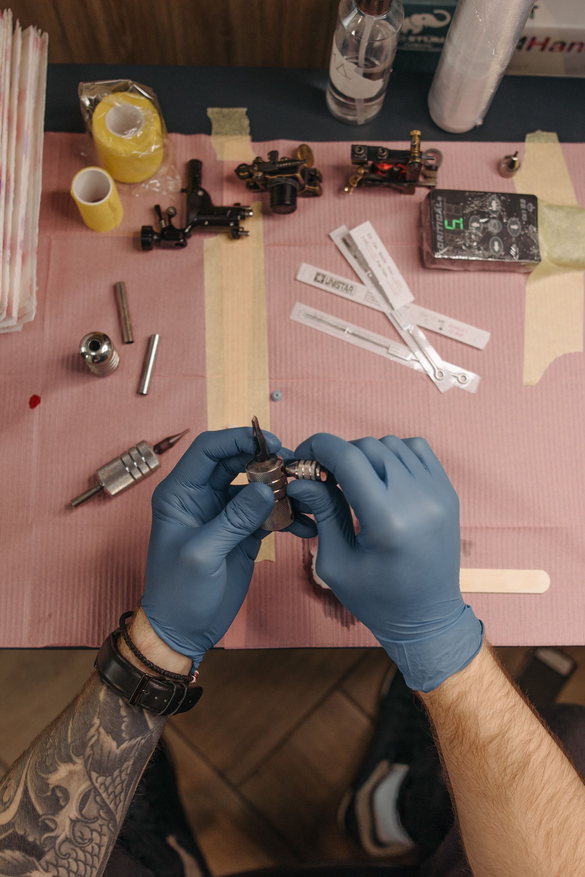 Top-down view of a tattoo workstation with machines, tools, supplies, packaging and blue-gloved hands handling equipment.