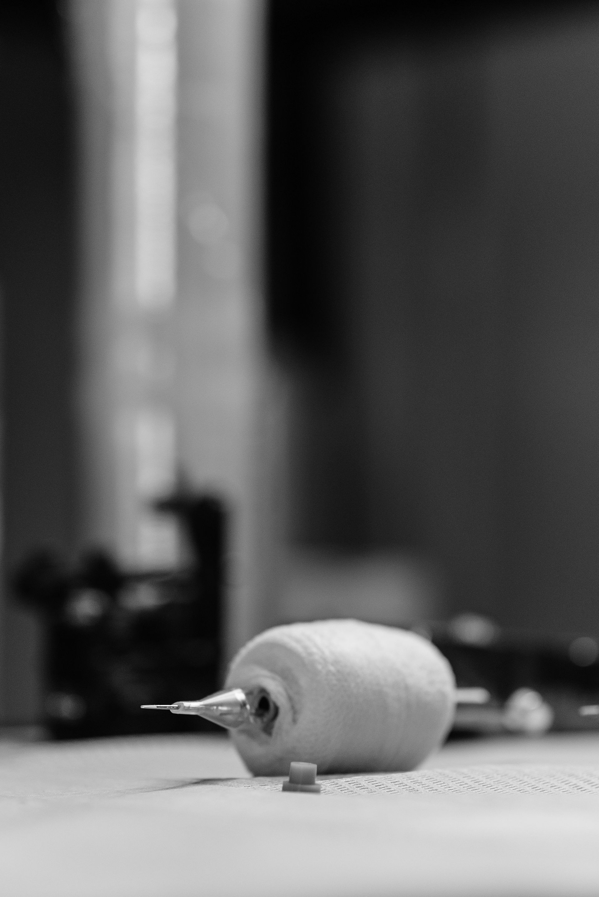 A monochrome close-up shows a tattoo needle cartridge and related equipment resting on a studio workstation.