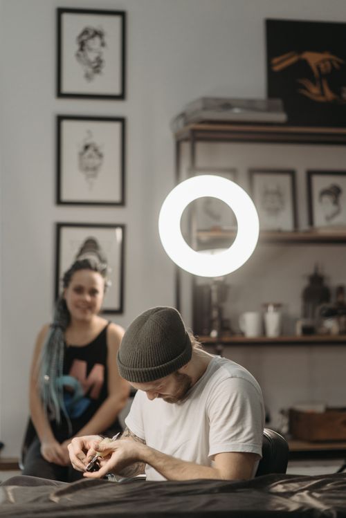 Free Tattoo artist focused on design in a modern studio setup with a curious observer. Stock Photo