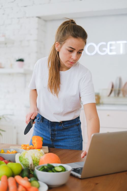 Free Young woman slicing vegetables while looking at a recipe on a laptop in a bright kitchen. Stock Photo
