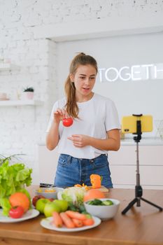 A woman records a culinary video with fresh vegetables using a smartphone and tripod in a bright kitchen.