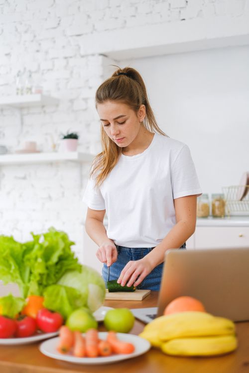 Free A young woman in a white t-shirt chops fresh vegetables on a wooden board in a bright kitchen. Stock Photo
