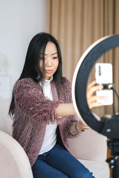 Asian woman using smartphone and ring light for selfie at home, showcasing modern content creation.