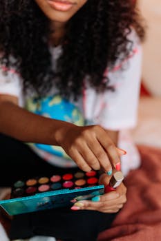 Close-up of a woman with curly hair using a vibrant eyeshadow palette and wearing bright nail polish.