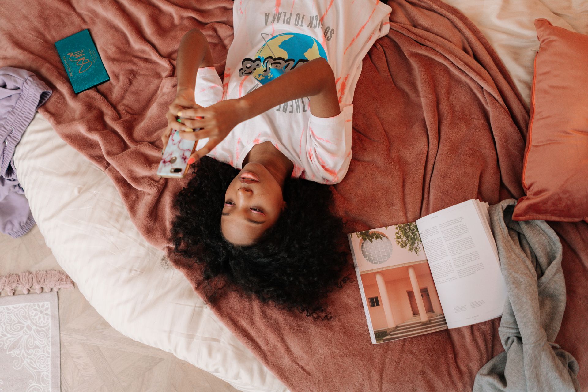 Young girl lying on bed, using smartphone and reading a book, with cozy blankets and soft pillows.