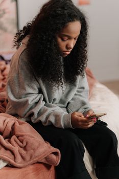 Teen girl with curly hair using smartphone while sitting on a bed indoors.