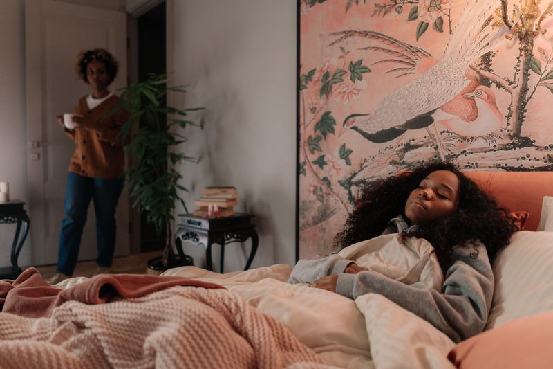 A Black mother carries a warm drink to her daughter, who rests in bed with her eyes closed. Past the baby red lines, fluids, rest, and a parent watching how the child looks and acts are the heart of home fever care.