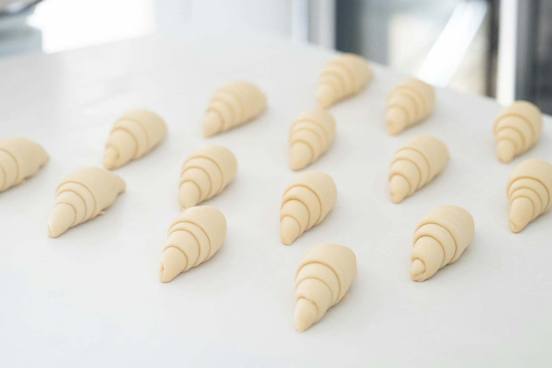Pre-baked croissant dough arranged on a baking sheet with parchment paper in a kitchen setting.