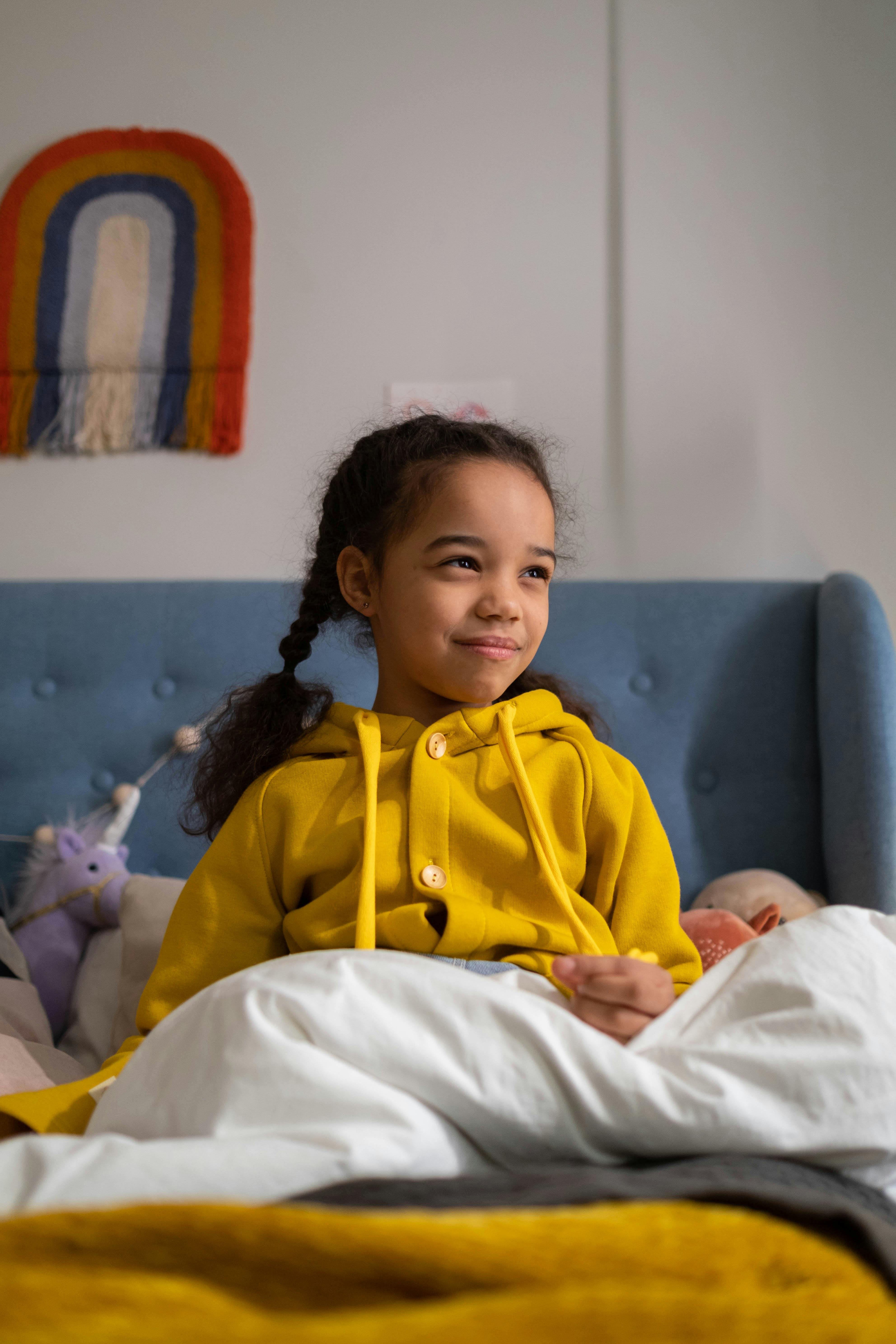 Smiling child in bedroom sitting upright in bed with a colorful wall hanging.
