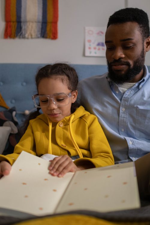 Free Father and daughter reading a book on a cozy bed in a warm indoor setting. Stock Photo