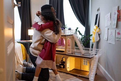 Free A father warmly embraces his daughter beside a dollhouse, capturing an affectionate moment indoors. Stock Photo