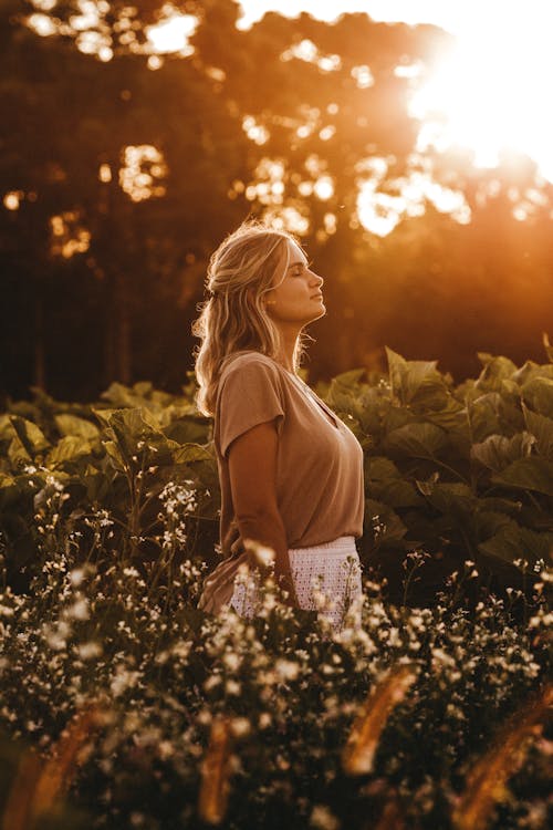 Seitenansicht einer attraktiven jungen Frau in lässiger Sommerkleidung, die bei Sonnenuntergang mit geschlossenen Augen auf einem üppigen Feld in einer friedlichen Landschaft steht