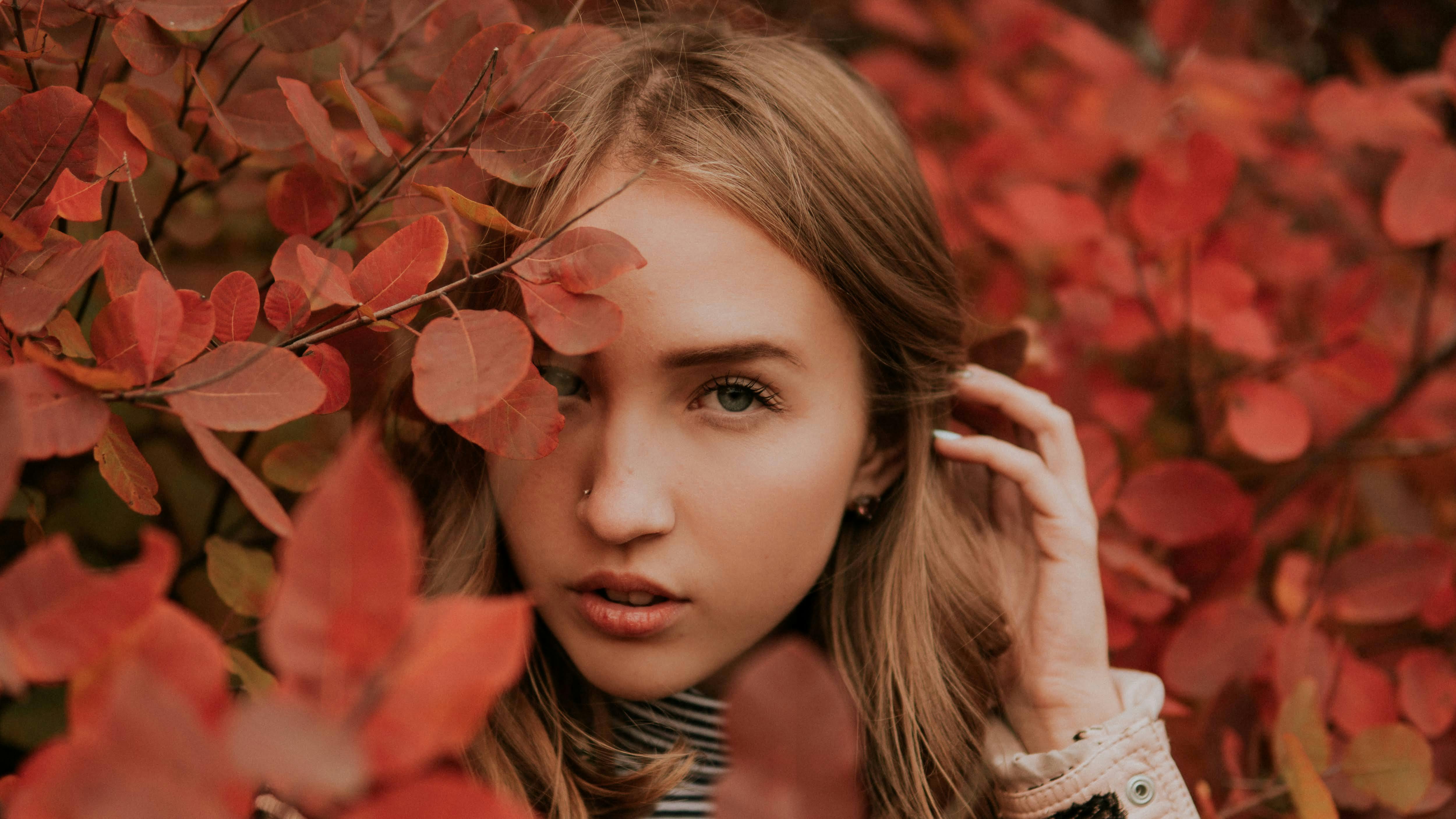 Serious young woman in warm clothes near shrubs and plants with red leaves and looking at camera in autumn day in nature