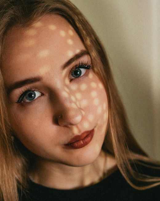 Thoughtful young lady with red lipstick and makeup with piercing in nose looking at camera with sunny dots on face on white background
