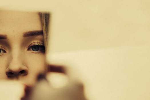 Unrecognizable crop young lady with makeup and piercing in nose looking in small mirror in light room