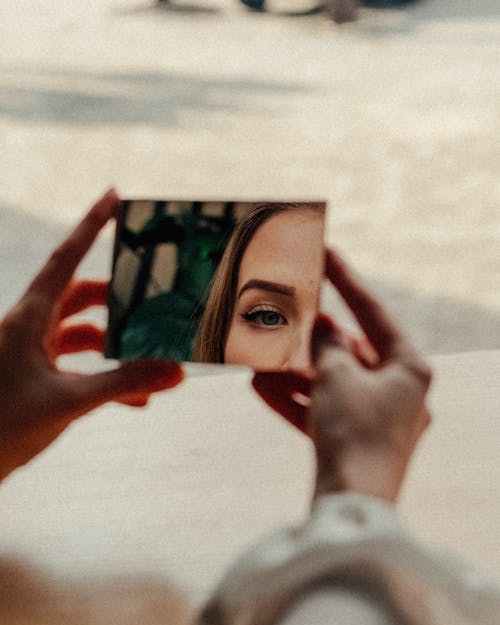 Faceless crop young lady with makeup looking in small mirror in hands in daylight in street