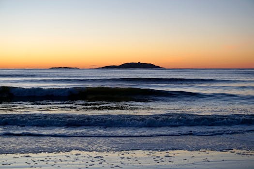 Tranquil view of ocean waves at sunrise, featuring a distant island under a glowing sky.