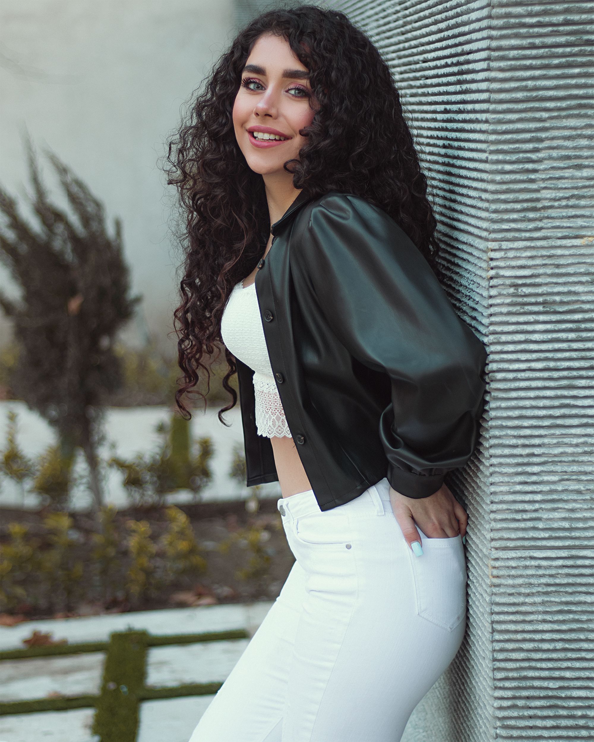 Stylish woman with curly hair posing outdoors in a black leather jacket and white pants.