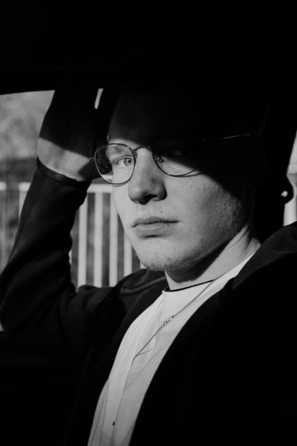 Black and white of calm young male in casual clothes and eyeglasses siting in car and looking at camera on sunny day
