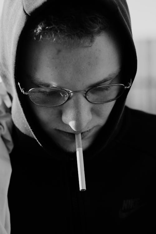 Black and white of thoughtful young male millennial in hooded sweatshirt and eyeglasses smoking cigarette on street on sunny day