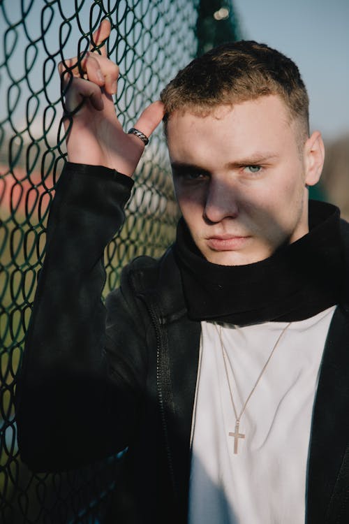 Focused young male millennial in casual clothes and accessories standing near chain link fence and looking at camera on sunny day in countryside