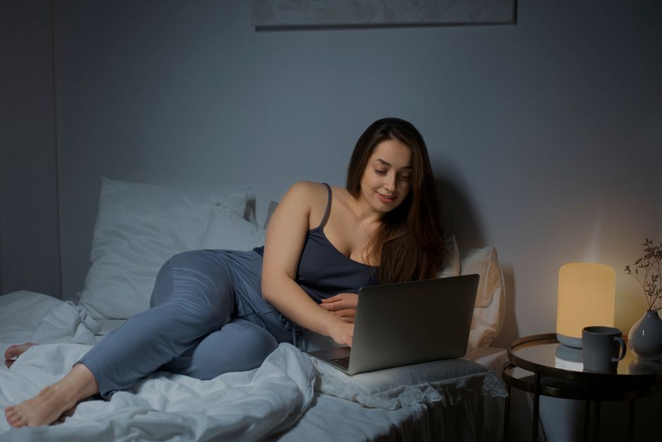 Woman lying in bed using a laptop, cozy setting with warm lighting.