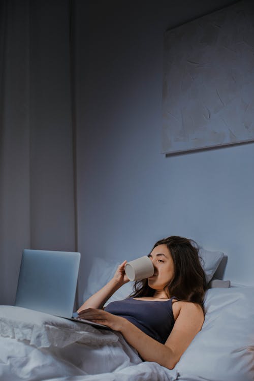 Free Woman in Blue Spaghetti Strap Top Reclining on Bed Using Laptop and Drinking Stock Photo