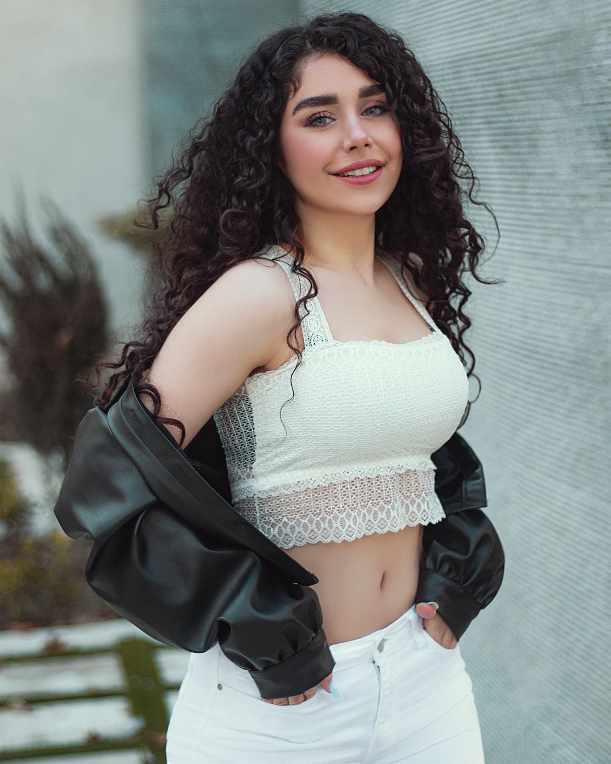 Young woman posing outdoors in a leather jacket and lace crop top, showcasing curly brunette hair and casual style.