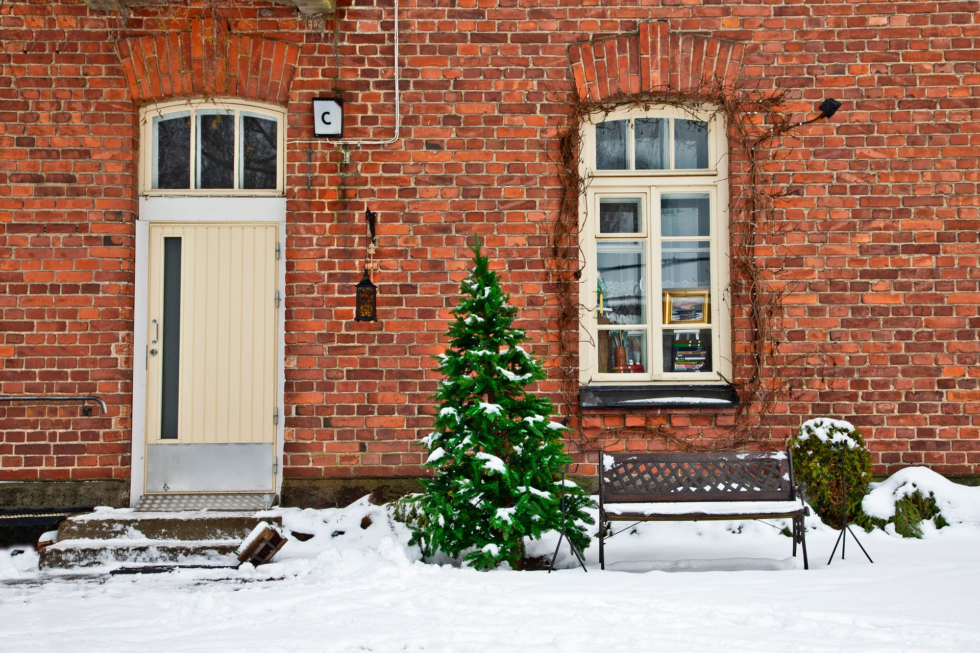 A cozy brick façade with snow-covered Christmas tree and bench beside a door and window in winter.