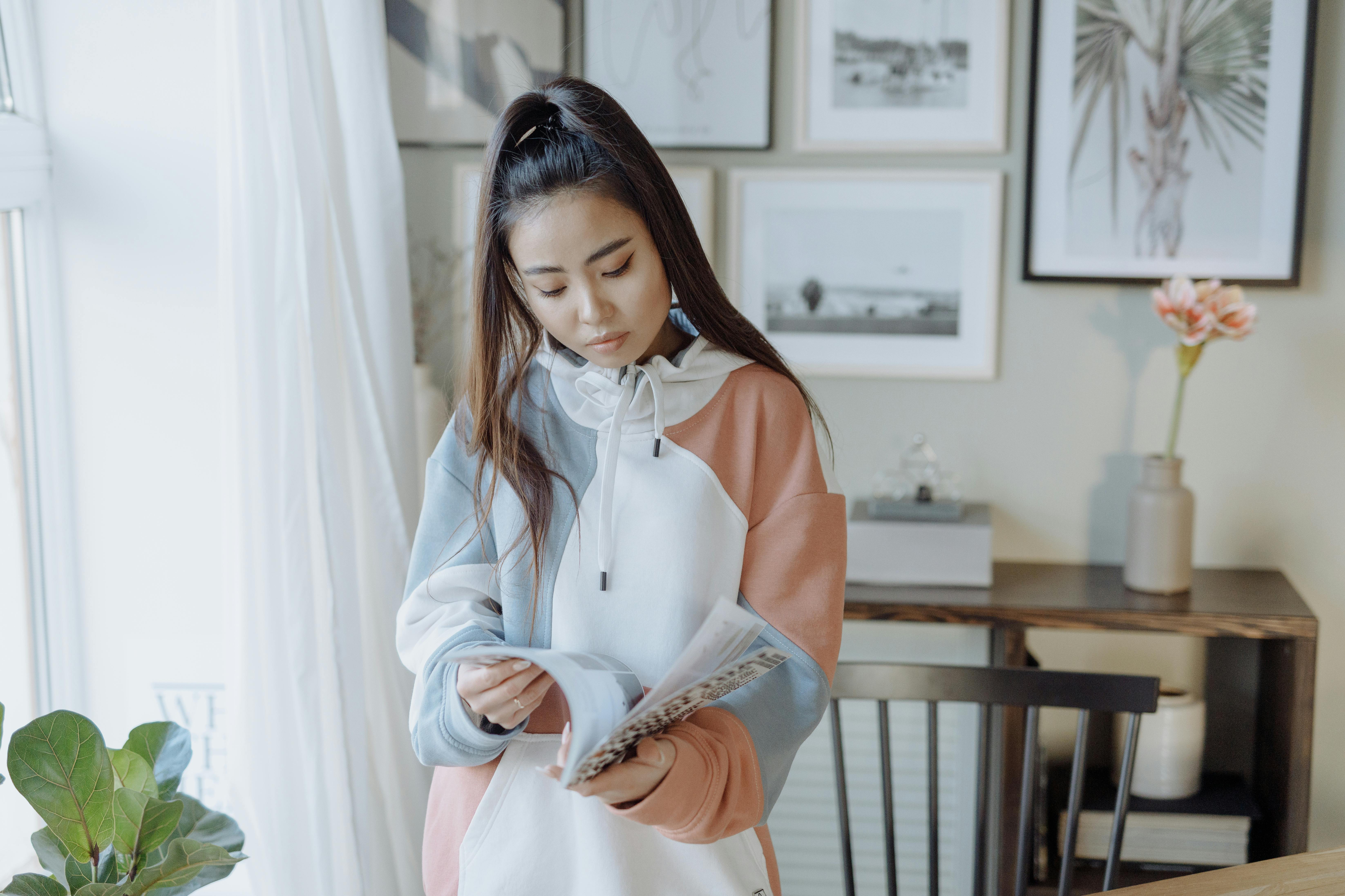Asian woman in a hoodie jacket reading a book in a peaceful home setting, surrounded by modern art.