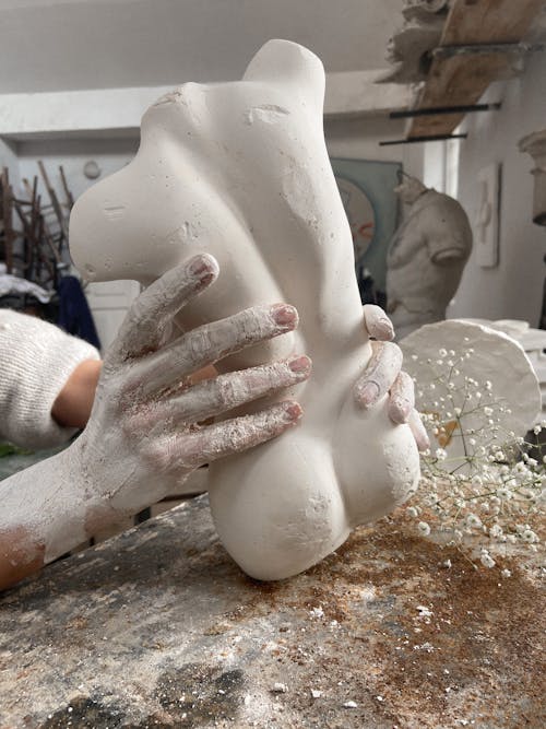 Unrecognizable artisan with dirty hands forming clay sculpture of torso while working at messy table with branch of flowers in professional workshop