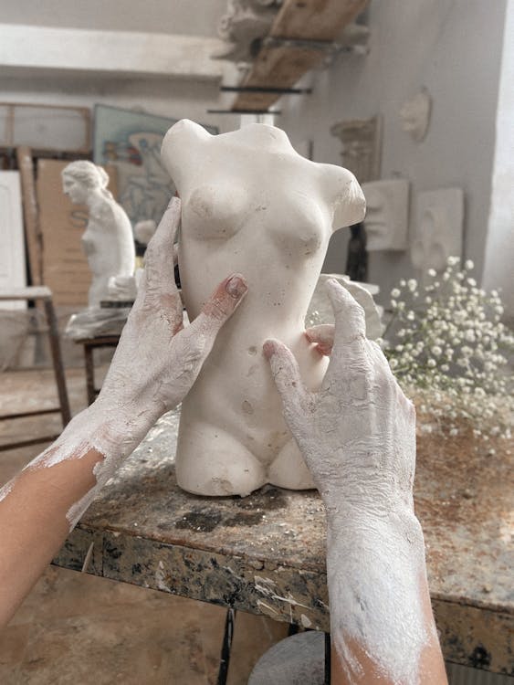 Free Unrecognizable talented artisan with dirty hands making clay female torso while sitting at wooden workbench with branch in modern workshop Stock Photo