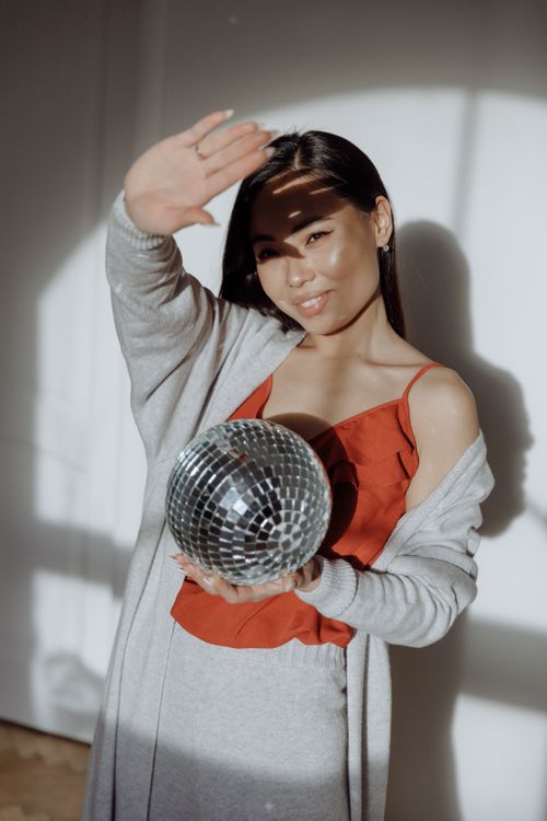 Free A young woman in a casual orange top holds a disco ball indoors, smiling warmly. Stock Photo