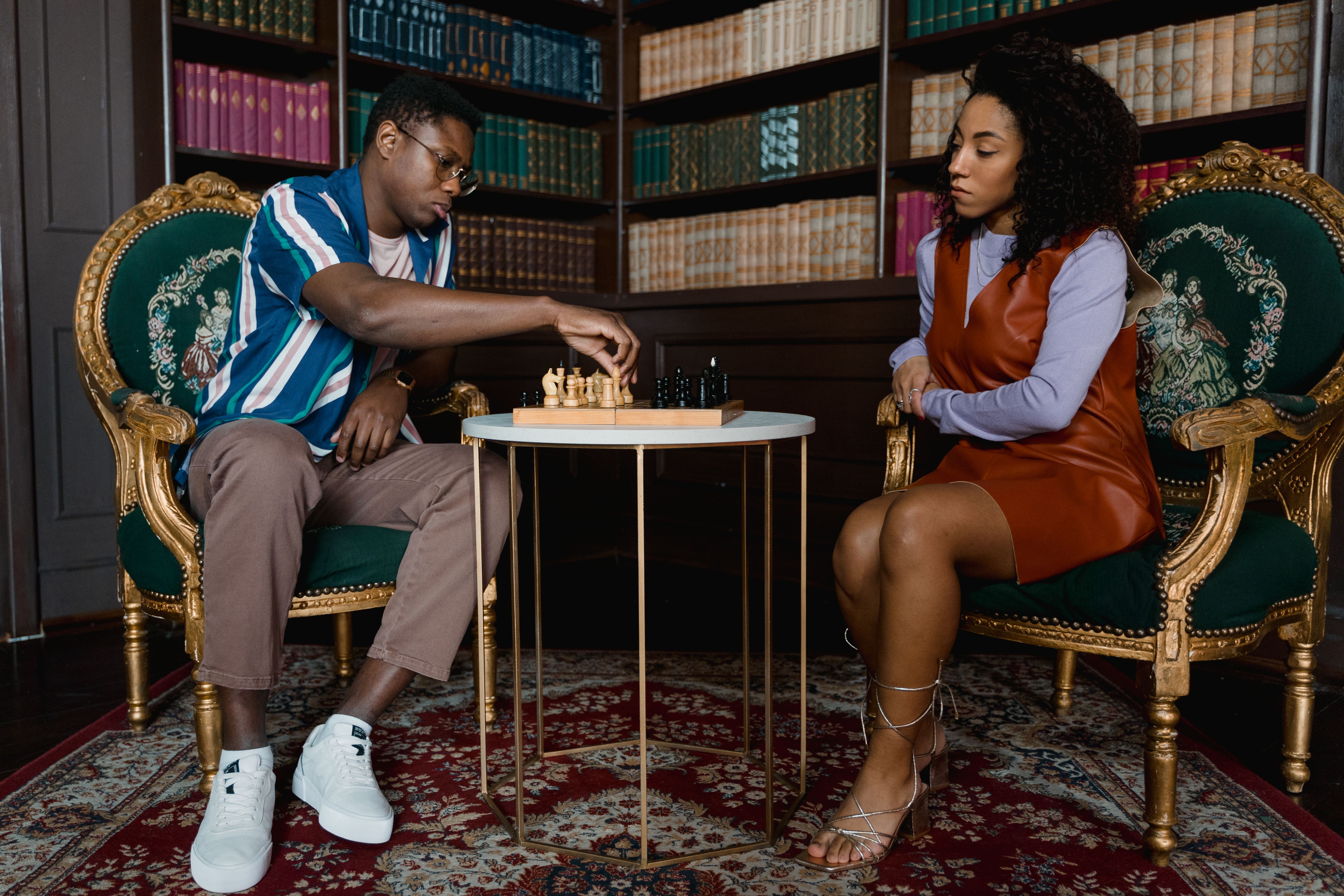 A couple intensely focused on a chess game, sitting in a luxurious library setting.