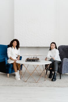 Two women seated in a modern room playing chess, showcasing focus and strategy.