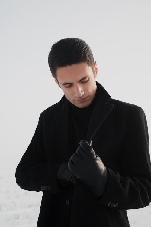 Confident young male with dark hair adjusting elegant black coat while standing on meadow covered with snow against overcast sky