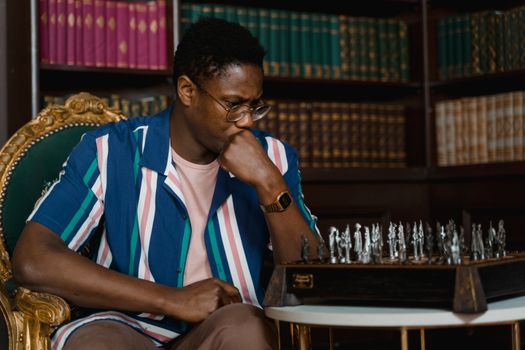 A man in a striped shirt concentrates on a chess game in an elegant library setting.