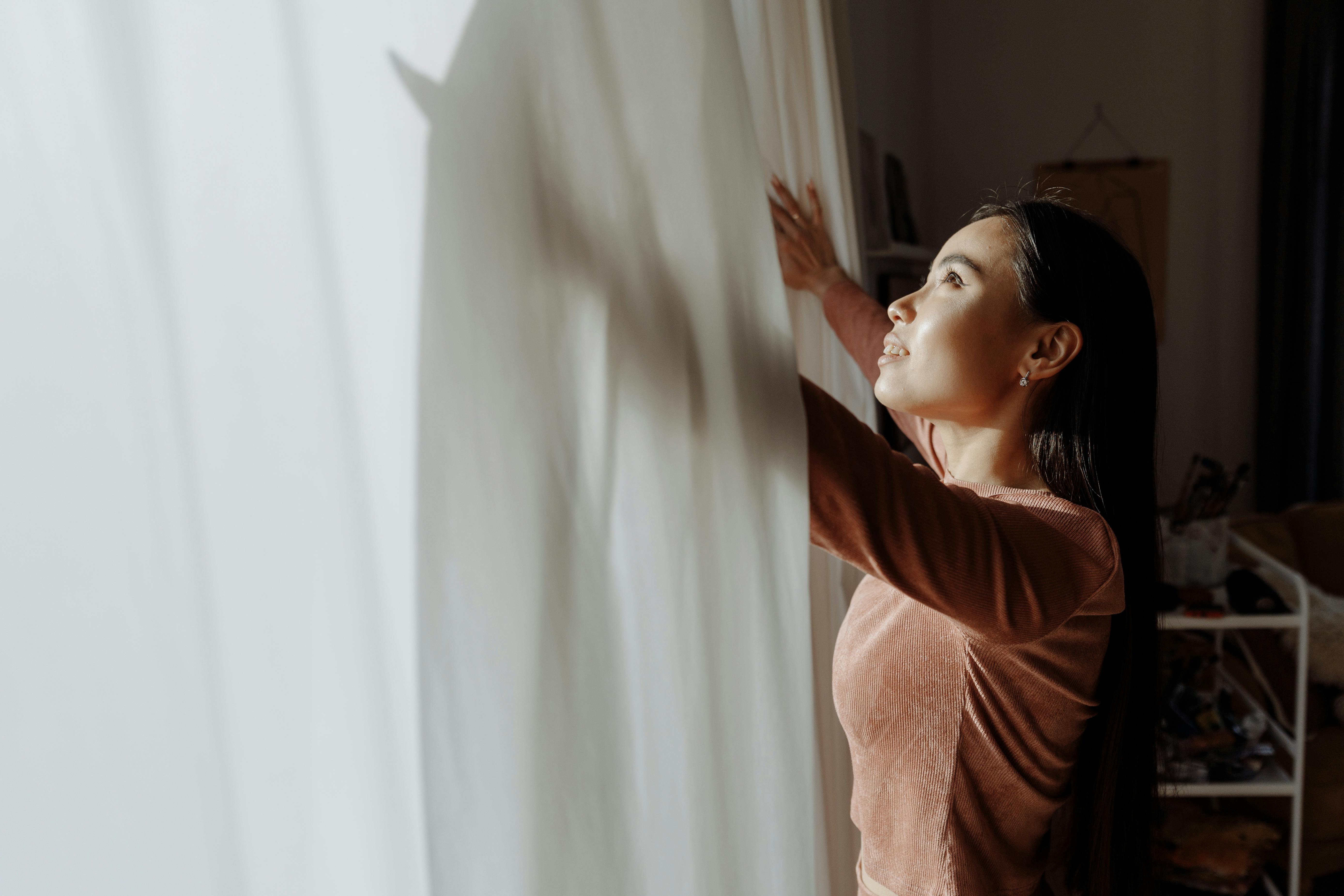 A woman embraces daylight near a white curtain, creating a serene indoor atmosphere.