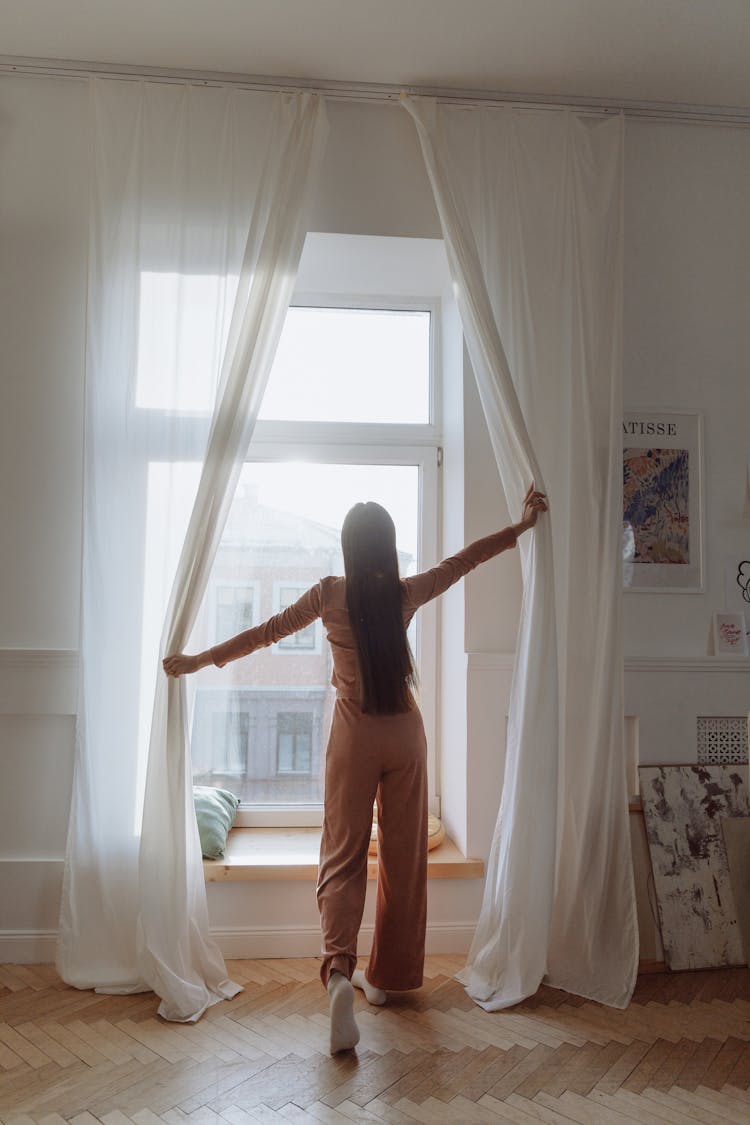 A Back View Of A Woman Standing Near The Window While Opening Curtains