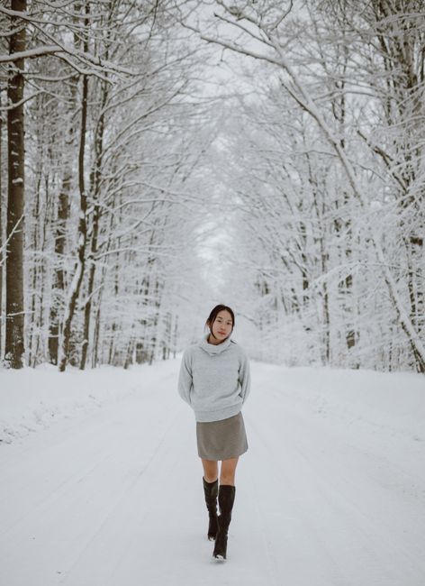 Full body of young slim Asian female with dark hair in stylish sweater and skirt walking on road covered with snow amidst leafless trees in winter