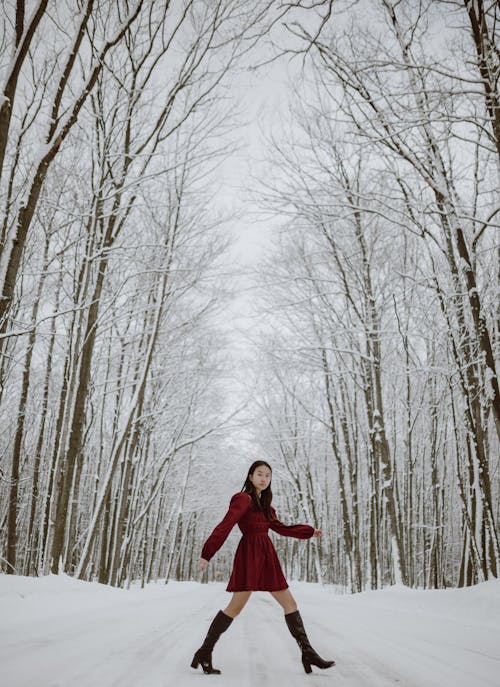 Side view full length of young gorgeous Asian female in red dress and boots walking on path covered with snow in woods with tall bare trees