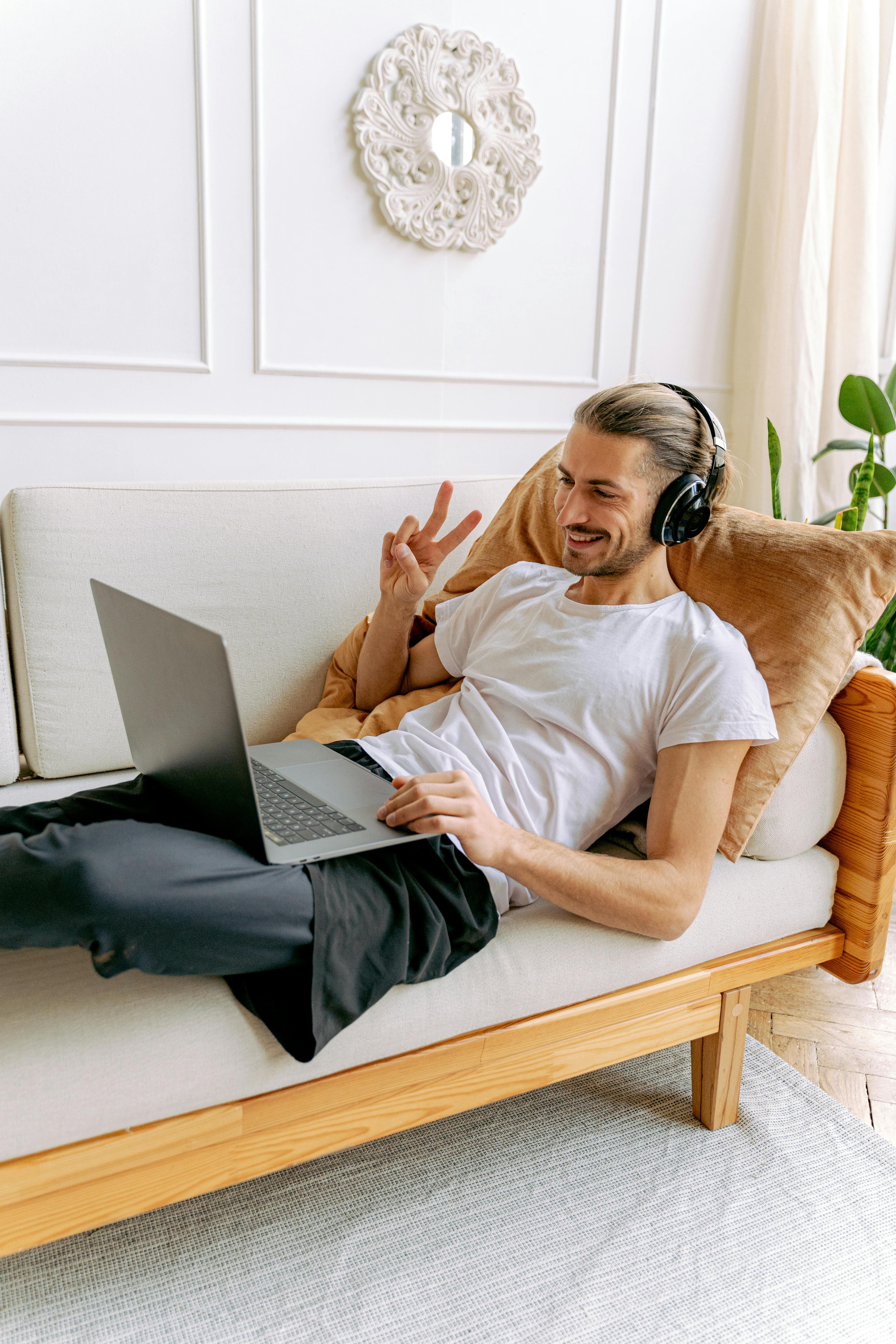 Free A man in a white shirt enjoys a relaxed moment with headphones and a laptop on a cozy sofa. Stock Photo