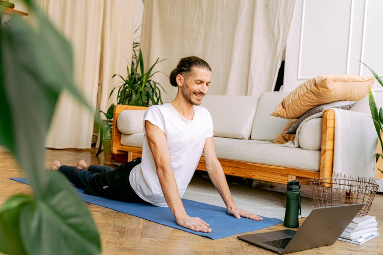 A Man In White Shirt Stretching His Body On The Floor While Looking At The Laptop