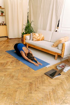 Woman doing yoga indoors on a mat with a laptop nearby, promoting wellness and fitness.