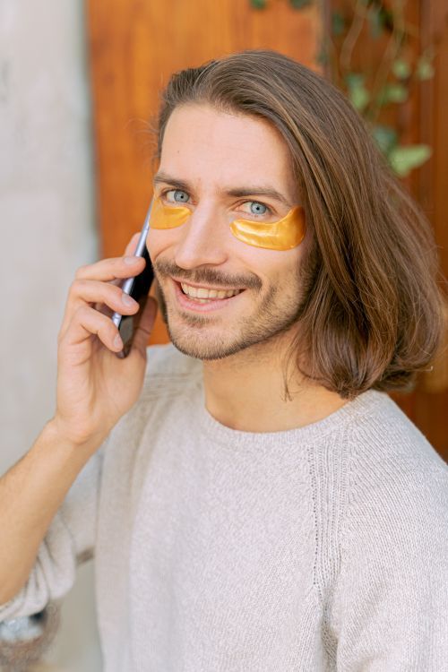 Free Smiling man with long hair using a phone, wearing under-eye patches outdoors. Stock Photo