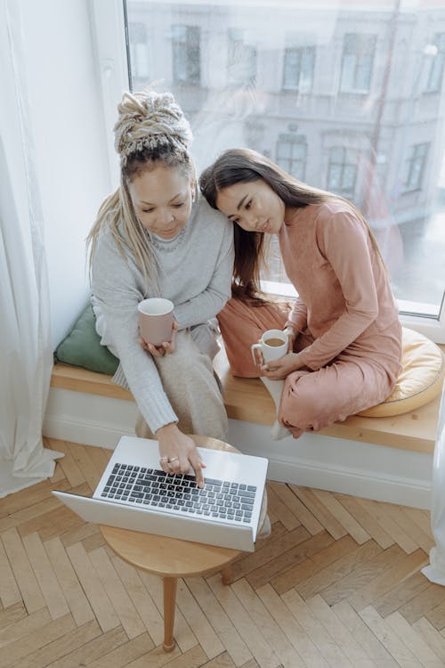 Free Women Sitting by the Glass Window while Using Laptop and Holding a Drink Stock Photo